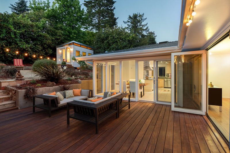 Backyard deck with dark wood seating and orange cushions, string lights, and glass sliding doors to a bright kitchen; hill and trees in back.