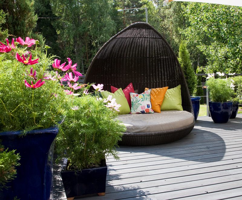 A dark wicker dome daybed with beige cushion and multicolored pillows on a wooden deck, surrounded by blue planters with pink flowers and green trees.