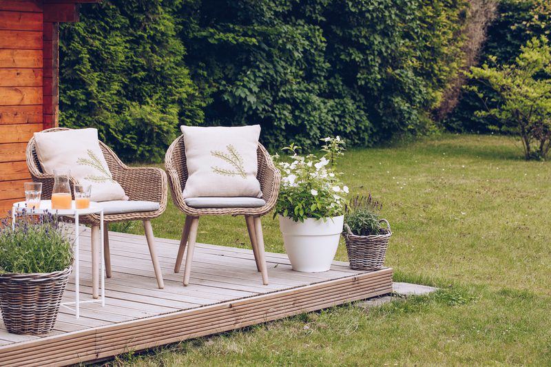 Wooden deck with two wicker chairs and cushions, a small white table with orange juice glasses, and potted plants beside a green lawn.
