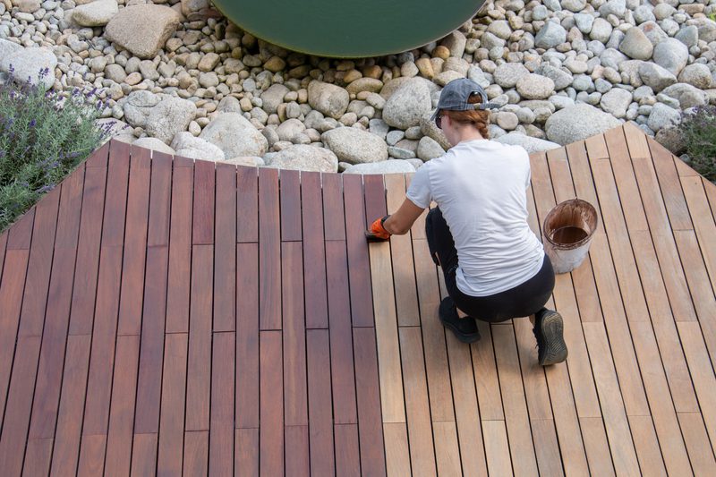 Person kneels on a wooden deck, applying finish with an orange-gloved hand; a bucket sits nearby as rocks and lavender border the area.
