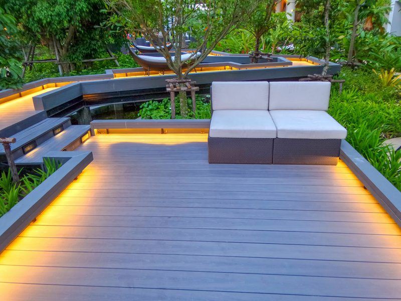 Modern outdoor deck with grey wicker sectional cushions, warm amber edge lighting, and lush greenery; multi-level platforms frame the seating.