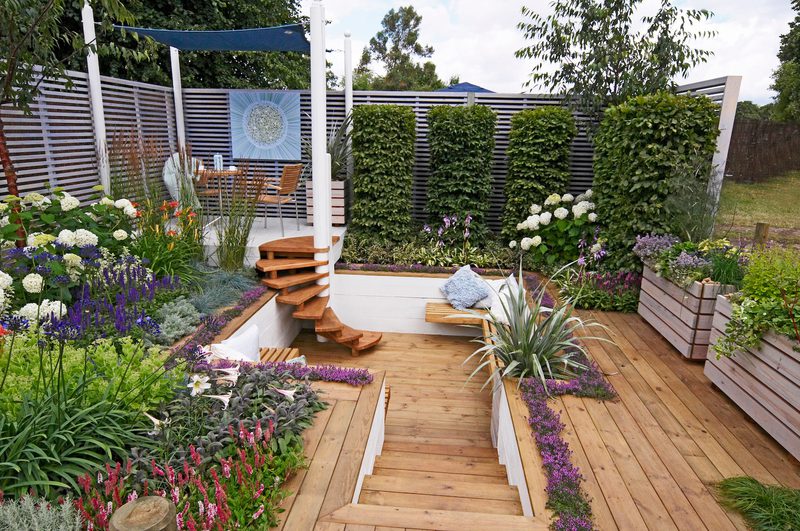Wooden deck garden with curved stairs, raised planters, purple and white flowers, tall hedges, and a shaded seating area with a blue art panel.