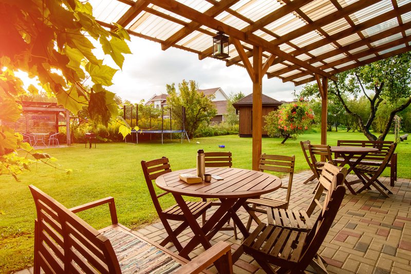 Sunlit backyard under a wooden pergola, round table with chairs on a brick patio, green lawn, a trampoline, trees, and hanging flowers.