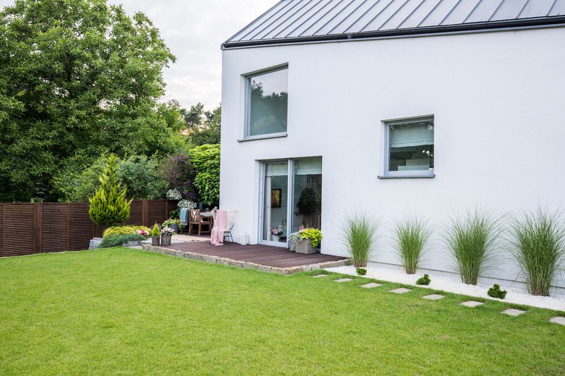 White modern house with a wooden deck and glass doors, potted plants, a green lawn, and tall grasses along the side wall; wooden fence on the left.