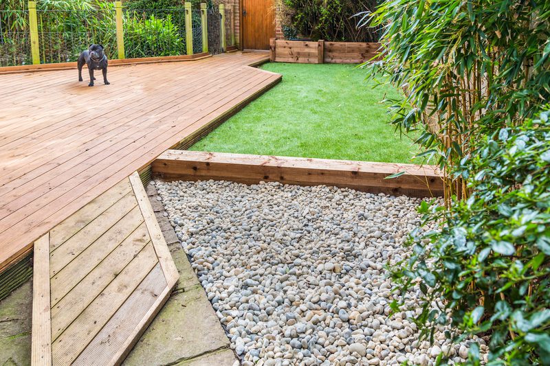 Backyard scene with a wooden deck, a small dark dog on the deck, a pebble area with wooden edging, and a green lawn bordered by plants.