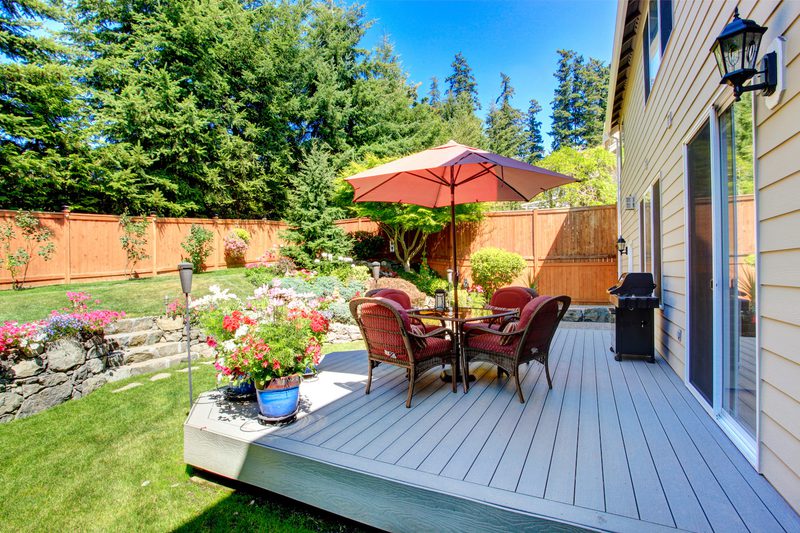 Backyard deck with a round table and six red-cushioned chairs under a pink umbrella, bordered by potted flowers, a grill, and a wooden privacy fence.