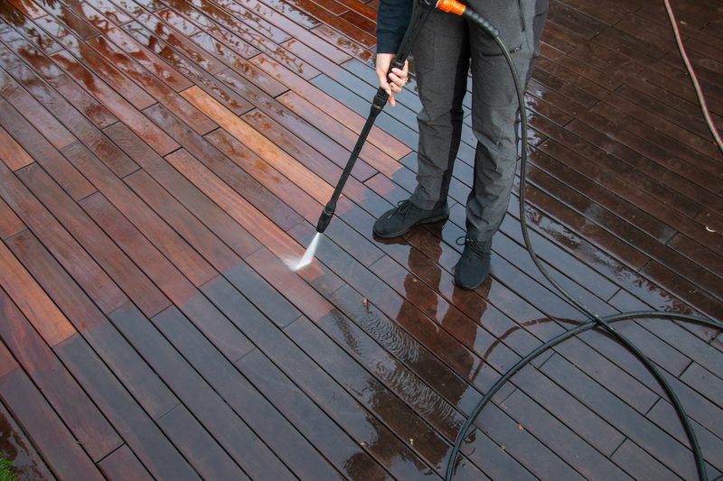 Person standing on a wet wooden deck, cleaning it with a pressure washer, holding a spray nozzle with a looping hose.