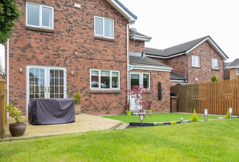 Backyard of a red brick two-story house with white-framed doors, a covered patio, potted plants, a small tree, artificial turf, and a wooden fence.