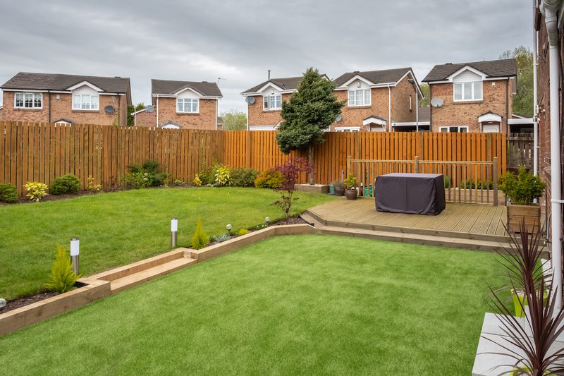 Backyard with a green lawn, wooden fence, and raised deck with a covered grill; potted plants along the deck and a small tree, brick houses beyond.