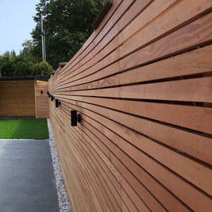 Long horizontal wooden slat fence with black outdoor lights, beside a paved path with a gravel border and a green lawn visible beyond.