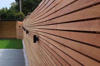 Long horizontal wooden slat fence with black outdoor lights, beside a paved path with a gravel border and a green lawn visible beyond.