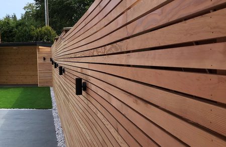 Long horizontal wooden slat fence with black outdoor lights, beside a paved path with a gravel border and a green lawn visible beyond.