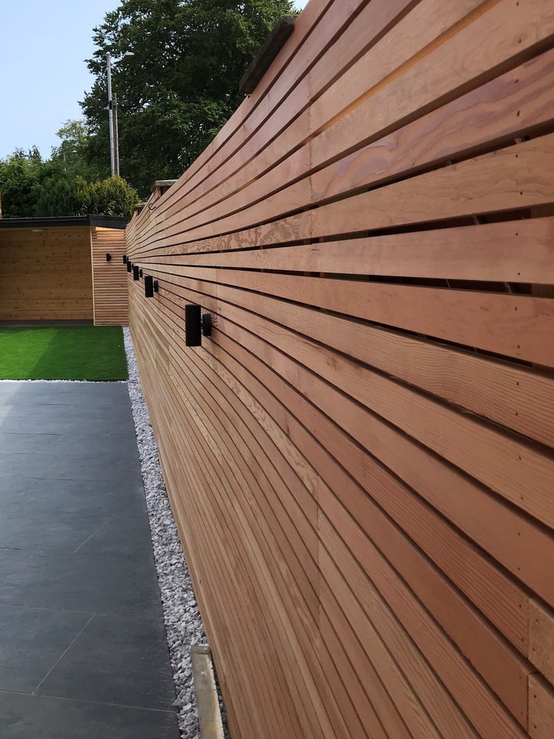 Long horizontal wooden slat fence with black outdoor lights, beside a paved path with a gravel border and a green lawn visible beyond.