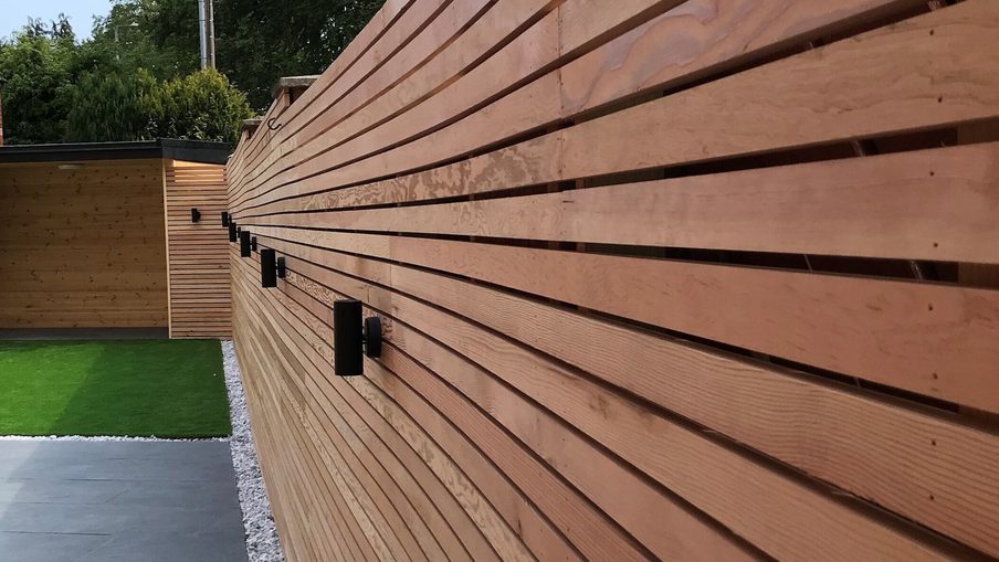 Long horizontal wooden slat fence with black outdoor lights, beside a paved path with a gravel border and a green lawn visible beyond.