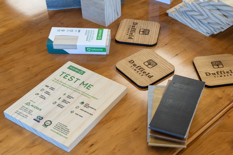 Wooden table with Accoya TEST ME board, Duffield Timber coasters, a small box, and stacked dark wood sample tiles.