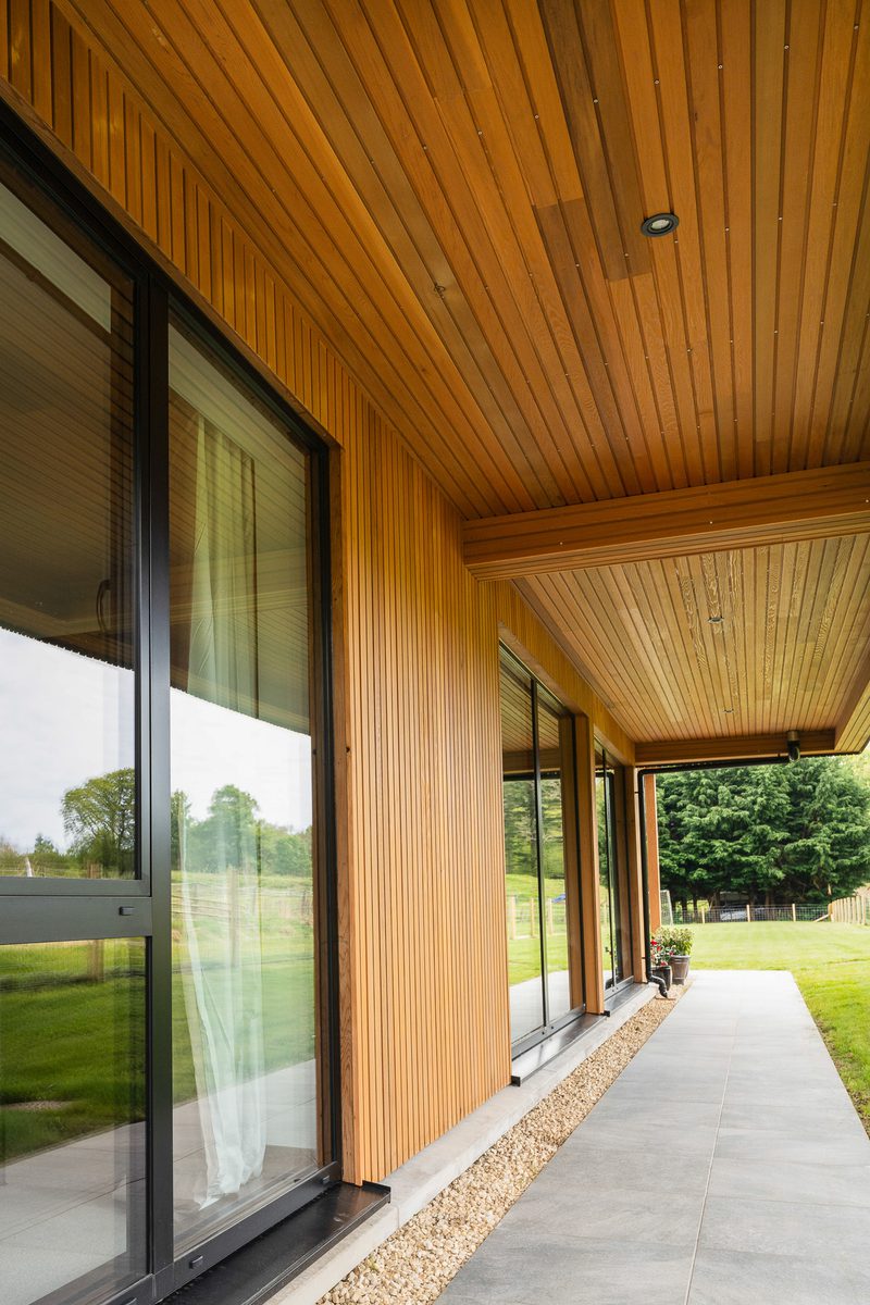Modern wooden-clad building with tall glass doors, a covered veranda, a gravel border, and a green lawn with trees in the background.
