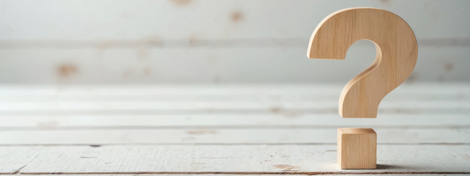 A wooden question-mark sculpture sits on a small square base on a pale, weathered wooden surface.