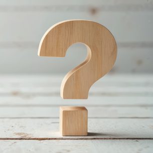 A 3D light wood question mark sculpture on a small square base rests on a pale, weathered wooden surface.