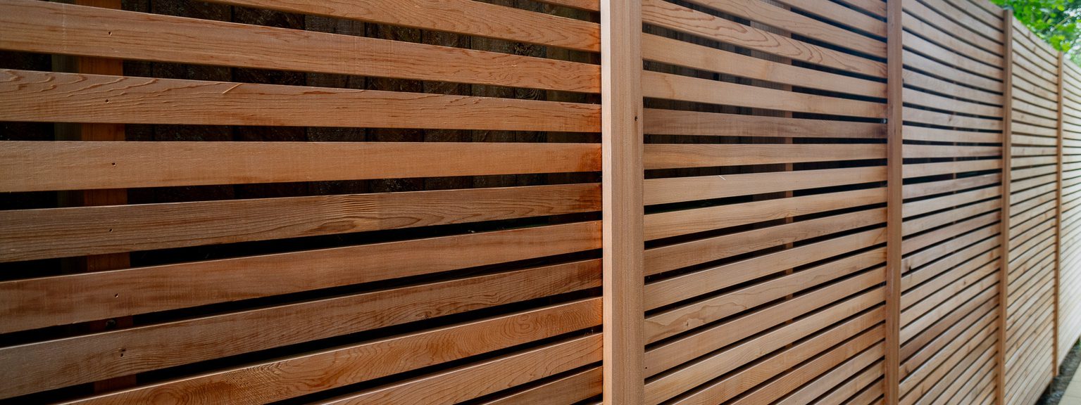 Long brown wooden fence with evenly spaced horizontal slats and vertical posts along a sidewalk, with green leaves visible above.