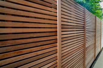 Long brown wooden fence with evenly spaced horizontal slats and vertical posts along a sidewalk, with green leaves visible above.