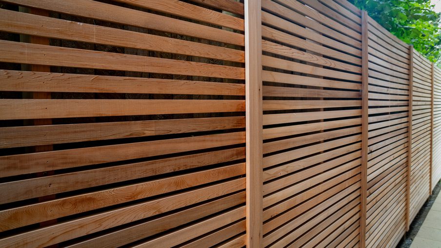 Long brown wooden fence with evenly spaced horizontal slats and vertical posts along a sidewalk, with green leaves visible above.