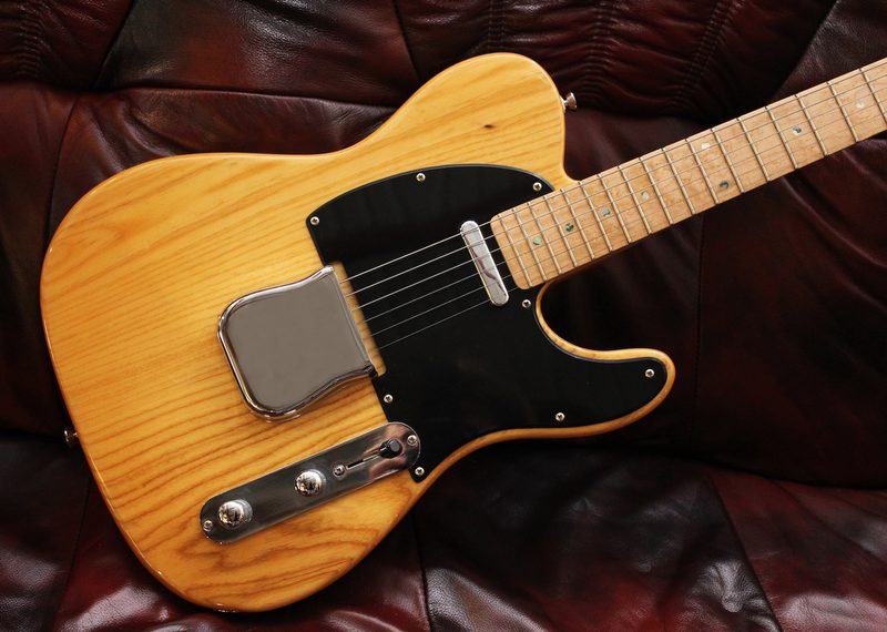 Natural-blonde electric guitar with a black pickguard and chrome hardware resting on a dark burgundy leather couch.
