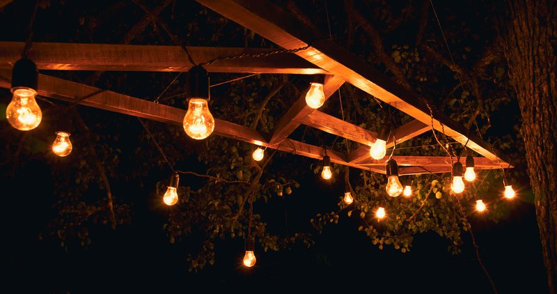String of warm incandescent bulbs hung from a wooden frame above, with tree branches in the background, at night, creating a cozy glow.