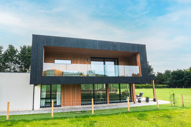 Contemporary two-story house with dark siding, wood accents, and a glass balcony; large windows overlook a green lawn, fence, and trees.