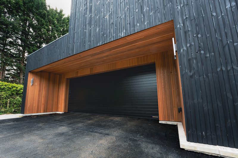 Modern black garage door set into a dark wooden-clad building with a warm wooden overhang; trees and greenery on the left and a paved driveway.