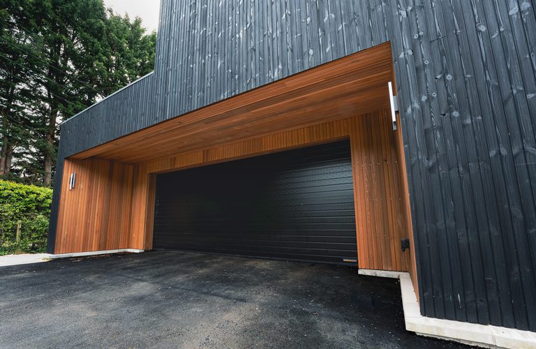 Modern building with dark vertical wood siding and a large black garage door framed by warm wood under a wooden overhang.