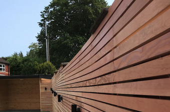 Long wooden slat fence runs along a yard, with a wooden shed to the left, brick houses in the background, and tall trees under a clear blue sky.