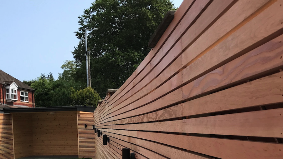 Long wooden slat fence runs along a yard, with a wooden shed to the left, brick houses in the background, and tall trees under a clear blue sky.