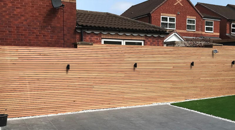 Horizontal wooden fence with black outdoor lights, in front of red brick houses. A gray patio with a white gravel border and a green lawn strip.