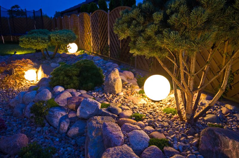 Night garden with rock and gravel ground, small shrubs and trees, illuminated by glowing spherical lamps beside a wooden lattice fence.