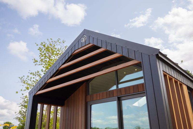Modern dark wooden tiny house with large glass windows, an angled roof with exposed wooden beams, and a blue-sky backdrop with trees.