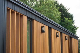 Modern exterior with vertical warm-brown wooden slats, black trim, and wall-mounted lanterns along a row of windows; green trees in the background.