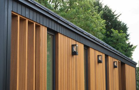 Modern exterior with vertical warm-brown wooden slats, black trim, and wall-mounted lanterns along a row of windows; green trees in the background.