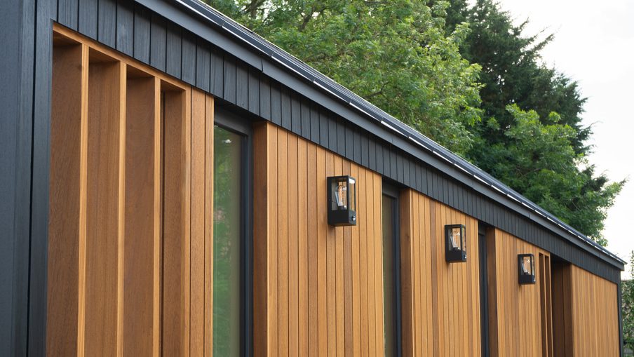 Modern exterior with vertical warm-brown wooden slats, black trim, and wall-mounted lanterns along a row of windows; green trees in the background.