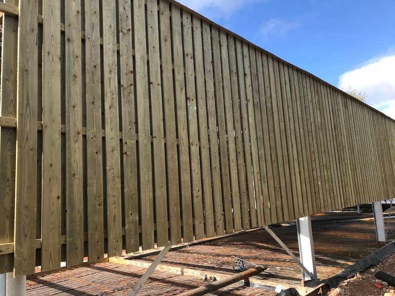 Tall wooden fence with vertical slats on metal supports, elevated above a brick-paved construction site beneath a blue sky.