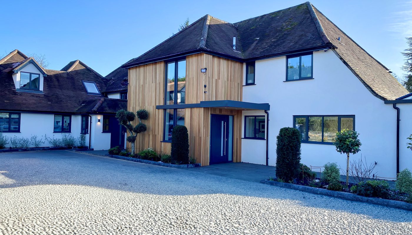Two-story modern house with white walls and wooden cladding at the entrance, blue front door, large windows, trimmed shrubs, and a pebble driveway on a sunny day.