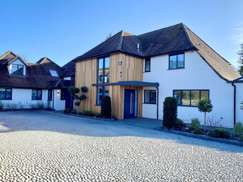 Two-story modern house with white walls and wooden cladding at the entrance, blue front door, large windows, trimmed shrubs, and a pebble driveway on a sunny day.