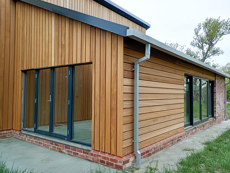 Modern wooden building with vertical panels and a brick base features large glass doors, gray gutter, and a paved path, with grass and trees in view.