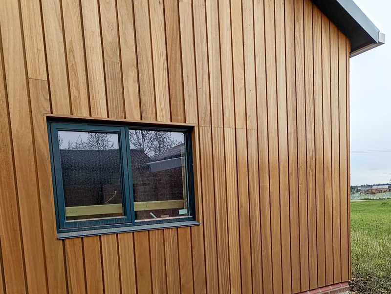 Brown vertical wooden siding with a blue-framed double window; reflections of trees in the glass, set on a grassy field under an overcast sky.