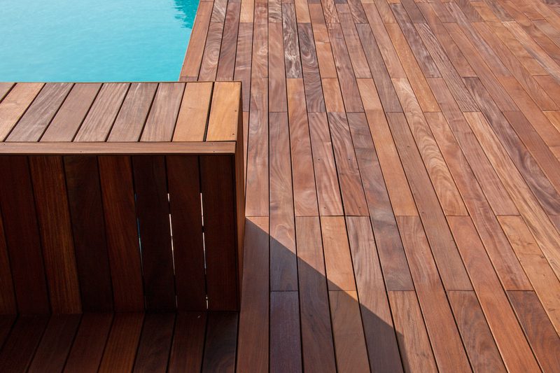 Sunlit wooden pool deck with long planks; a slatted wooden bench/railing on the left and a blue pool edge at the top, casting a shadow.