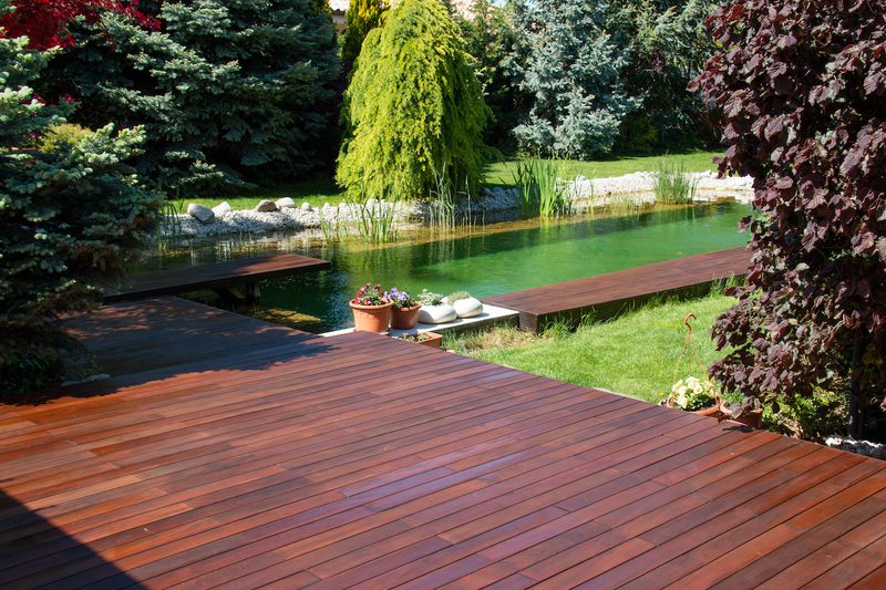 Wooden deck in the foreground overlooks a green, rocky-bordered pond; potted flowers rest on a small platform by the water.