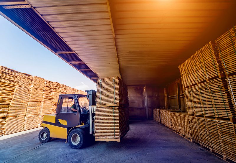 Yellow forklift with operator lifts a stack of wooden pallets inside a warehouse, flanked by tall pallet racks under a long metal roof.