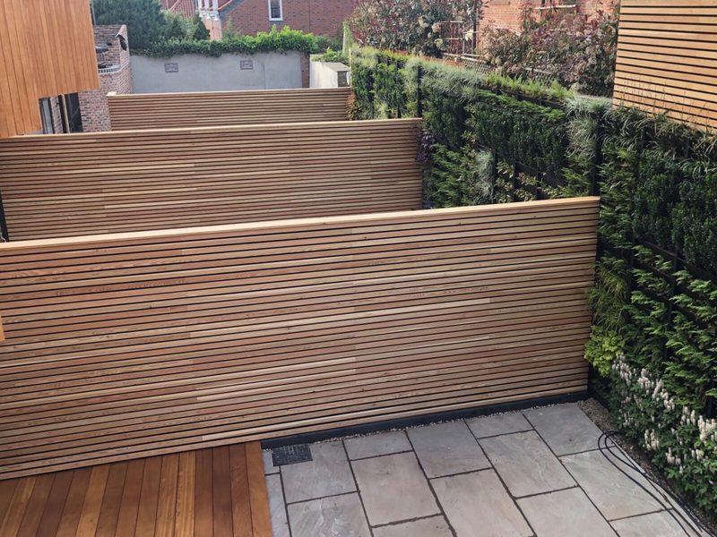 Backyard or patio area with horizontal wooden slat privacy screens, a light stone tile patio, and tall green hedges along a wooden fence.