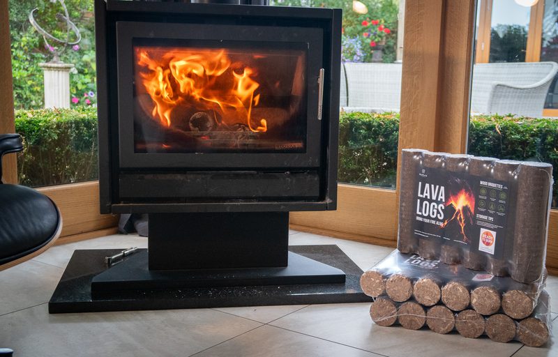 Black freestanding stove with a glass door showing flames; sits on a raised platform in a bright room with a stack of lava logs to the right.