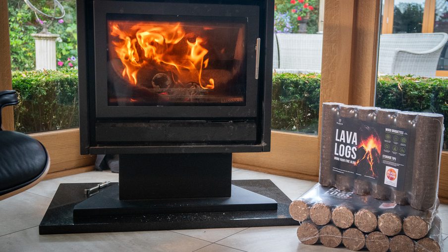 Black freestanding stove with a glass door showing flames; sits on a raised platform in a bright room with a stack of lava logs to the right.