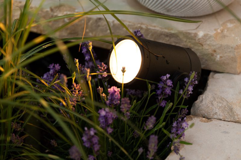 Black cylindrical outdoor spotlight glows warm white, nestled among purple lavender and green grasses in a stone-edged garden bed.